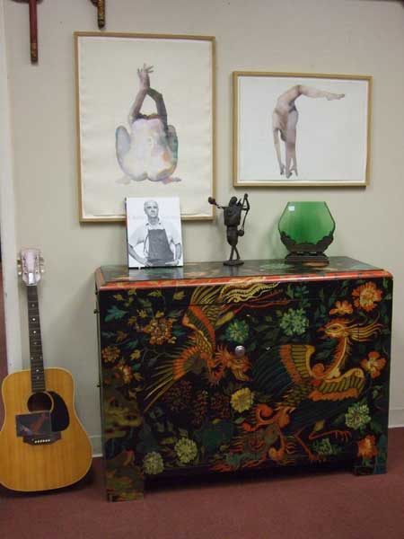 Ornate dresser with small art objects on top. Art drawings on the wall and a guitar beside dresser.